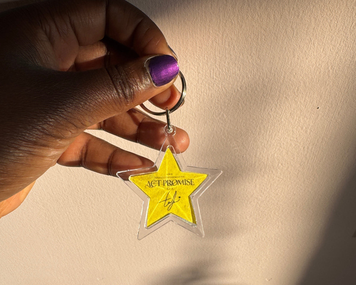 A hand with purple nail polish holds a clear star-shaped acrylic keychain with a bright yellow center featuring the text 'Act Promise' and a signature, set against a wall with dramatic sunlight and shadows.